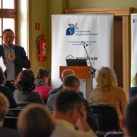 Konferencja. Uczestnicy spotkania siedzą na krzesłach, jeden z prelegentów wygłasza prezentację